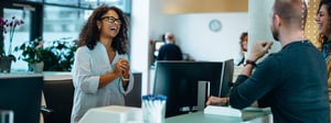 A receptionist greeting an expected guest at an office front desk, with the visit already registered against the meeting